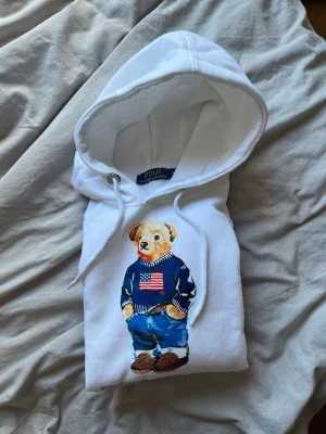 Vit hoodie med Polo Bear från Polo Ralph Lauren - Vit hoodie från Polo Ralph Lauren med den ikoniska Polo Bear-trycket framtill. Björnen har på sig en blå tröja med amerikanska flaggan och jeans. Hoodien har dragsko i huvan och är gjord i mjukt material. Perfekt för en avslappnad och cool stil.