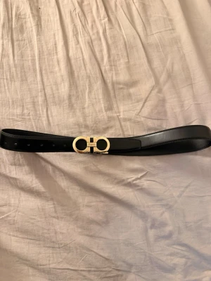 Svart skärp från Ferragamo - Snyggt svart skärp från Ferragamo med ikoniskt guldigt spänne i metall. Skärpet är stilrent och har en klassisk design som passar till många olika outfits. Tillverkat i slätt skinn och har justerbara hål för perfekt passform.