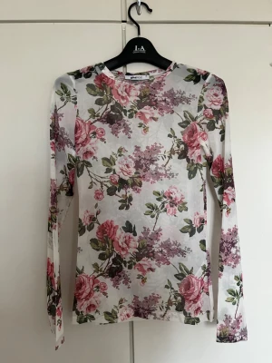 Blommig meshtröja från Gina Tricot - Säljer en långärmad meshtröja från Gina Tricot med blommönster i rosa, lila och grönt på vit botten. Tröjan har rund halsringning och är gjord i ett tunt, stretchigt material som känns lätt och luftigt.
