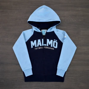 Malmö FF Hoodie Med Dragkedja  - Malmö FF hoodie med dragkedja — storlek 134/140 — NYSKICK Superfin hoodie från Malmö FF i marinblå och ljusblå färg med tryck “MALMÖ Fotboll Förening” ⚽🩵 ✔ Storlek: 134/140 ✔ Skick: Nyskick — knappt använd ✔ Dragkedja fram och huva ✔ Mjuk och skön kvalitet ✔ Officiell supportertröja ✔ Perfekt till skola, fritid eller match Från rök- och djurfritt hem.