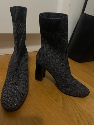 Glittriga svarta sockboots med klack - Säljer ett par svarta sockboots med glittrig finish och ribbad skaftkant. Bootsens material är stretchigt stickat tyg som sitter tight runt benet och har en fyrkantig tå samt bred hög klack. Perfekta för dig som vill ha en trendig och bekväm look.