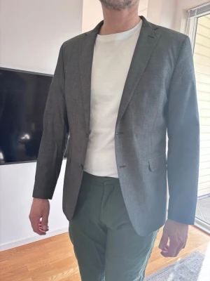 Mörkgrå klassisk kavaj herr - Stilren mörkgrå kavaj med smal passform och två knappar framtill. Klassisk design med spetsiga slag och två fickor med lock. Perfekt att matcha med både t-shirt och skjorta för en clean look. Materialet ser ut att vara en blandning av bomull och polyester.