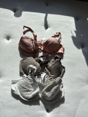 Spetsbhar i rosa, beige och vitt - Säljer tre bhar med vacker spetsdetalj i färgerna ljusrosa, beige och vit. Alla har justerbara axelband och bygel för extra stöd. Perfekta för dig som gillar romantisk och feminin stil. Märke och material framgår ej.