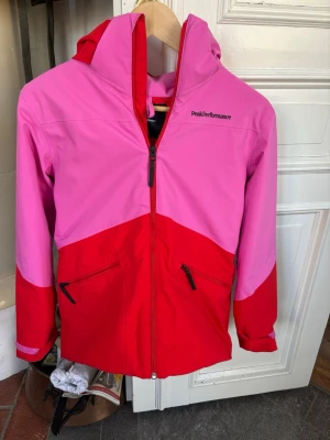 Peak Performance rosa/röd jacka  - Snygg jacka från Peak Performance i tvåfärgad design med rosa upptill och röd nedtill. Jackan har huva, hel dragkedja framtill och två snedställda fickor med dragkedja. Materialet är lätt och vindtätt, perfekt för blåsiga dagar.