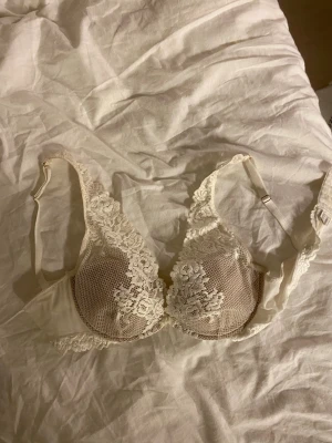 Vit spets-bh med vadderade kupor - Säljer en elegant vit bh med vadderade kupor och vackra spetsdetaljer framtill och på axelbanden. Bh:n har justerbara axelband och hakknäppning bak. Materialet är en kombination av spets och mjuk textil för extra komfort.