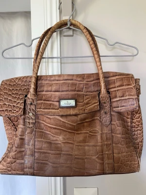 Brun handväska i krokopräglat läder - Snygg brun handväska från Tiger of Sweden i krokopräglat konstläder. Väskan har två rejäla handtag och en klassisk, rymlig design med lock och metallbricka framtill. 
