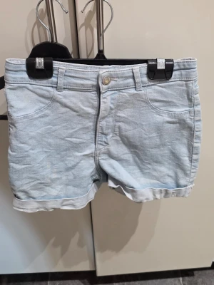 Ljusblå denimshorts med uppvik - Snygga ljusblå denimshorts med klassisk femficksdesign och uppvikta benslut. Shortsen har hällor för bälte och stängs med knapp och dragkedja. Perfekta för varma dagar och enkel att matcha med olika toppar.