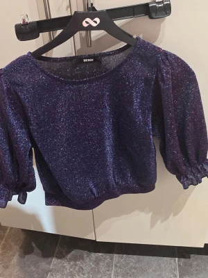 Glittrig lila topp från Bik Bok - Superfin glittrig lila topp från Bik Bok med puffiga trekvartsärmar och resår vid ärmslut och midja. Perfekt för dig som vill sticka ut med lite extra glitter och volym i outfiten.