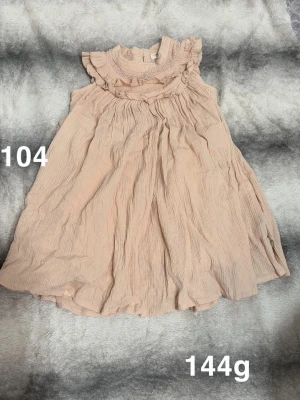 Aprikosfärgad volangklänning med hög krage - Söt aprikosfärgad klänning med volangdetaljer vid halsringningen och ärmlös design. Klänningen har en lätt, luftig känsla och är perfekt för varmare dagar.🌸 Fint använt skick. Storlek 104