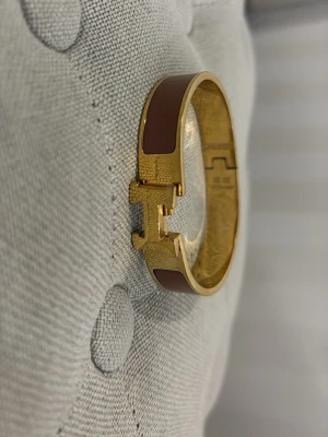 Ett H armband i brun  - Snyggt H armband i guldpläterad metall med bruna emaljdetaljer. Armbandet har den ikoniska H-formade låset och en stilren, rundad design som ger en lyxig känsla. Perfekt accessoar för att lyfta din outfit.