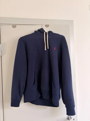 Marinblå hoodie från Polo Ralph Lauren - Snygg marinblå hoodie från Polo Ralph Lauren med klassisk huva och vita dragsnören. Har en röd broderad logga på bröstet och en stor magficka framtill. Perfekt för en avslappnad och stilren look.