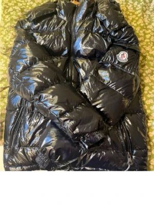 Svart glansig dunjacka från Moncler - Säljer en svart glansig dunjacka från Moncler med klassisk logotyp på ärmen. Jackan har dragkedja framtill, två sidofickor med dragkedja och en ficka på ärmen. Perfekt för kalla vinterdagar och riktigt snygg streetstil.