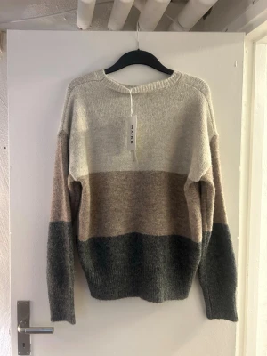 Randig stickad tröja från NA-KD - Mysig stickad tröja från NA-KD med breda blockränder i beige, brun och mörkgrå. Tröjan har rund halsringning, långa ärmar och avslappnad passform. Perfekt för lager-på-lager och chill dagar.