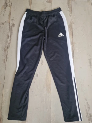 Svarta Adidas träningsbyxor - Snygga svarta träningsbyxor från Adidas med vita ränder längs sidorna och logga på låret. Byxorna har smal passform, resår i midjan och dragkedja vid benslut. Perfekta för träning eller sportiga outfits.