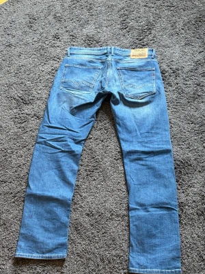 Blå raka jeans från Replay - Säljer ett par klassiska blå jeans från Replay med rak passform (Rocco). Jeansen har fem fickor, ljus tvätt och subtila slitningar framtill. Tillverkade i bomull med Nudie Jeans logga på bakfickan. Perfekta för en avslappnad och stilren look. Näst intill oanvända. Nypris 1499kr.
