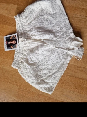 Vita spets-shorts med tofsar - Säljer ett par vita shorts med vacker spets och dekorativa tofsar framtill. Shortsen har elastisk midja och är perfekta för sommarens alla äventyr. De har en loose passform och känns både luftiga och stilrena.