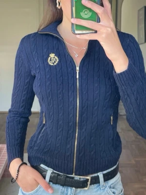 Mörkblå kabelstickad kofta Ralph lauren - Mörkblå kabelstickad kofta från Lauren Ralph Lauren med hel dragkedja, ståkrage och guldfärgade detaljer. Broderad logga på bröstet och två fickor med dragkedja. Perfekt för en snygg och klassisk look.