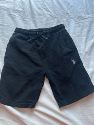 Svarta shorts från Polo Ralph Lauren - Svarta shorts från Polo Ralph Lauren med broderad logga på benet. De har elastisk midja med snörning och är tillverkade i mjuk bomull, perfekta för en avslappnad stil. För barn som är mellan 9-10 år.