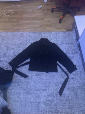 Svart trenchcoat från Gina Tricot - Snygg svart trenchcoat från Gina Tricot med klassisk dubbelknäppning och bälte i midjan. Jackan har långa ärmar och axelklaffar för en stilren look. Perfekt för dig som vill ha en tidlös och trendig jacka till garderoben.