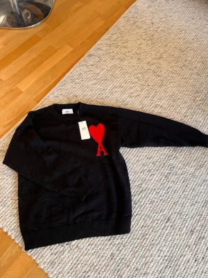 Svart Ami Paris sweatshirt med rött hjärta - Cool svart sweatshirt från Ami Paris med stort rött hjärta och bokstaven A broderat på bröstet. Tröjan har rund halsringning, ribbade muddar och är i mjukt material. Perfekt för en trendig och avslappnad stil.
