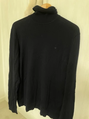 Svart polotröja från Calvin Klein - Svart stickad polotröja från Calvin Klein med diskret CK-logga på bröstet. Tröjan har lång ärm och klassisk polokrage, perfekt för lager-på-lager. Enkel och stilren design som funkar till det mesta. 100% Merino 