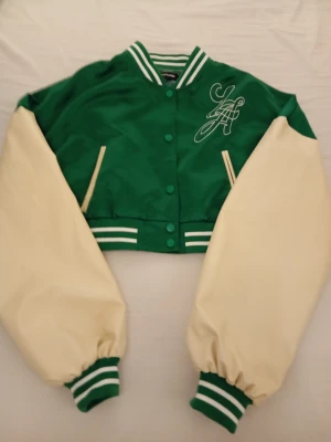 Grön och vit cropped bomberjacka - Cropped bomberjacka i grönt och vitt med broderad text 'Los Angeles' på ryggen och initialer fram. Jackan har gröna och vita ränder på muddar och krage, samt två snedställda fickor. Oversized ärmar i vitt och kropp i grönt ger en klassisk varsity-look.