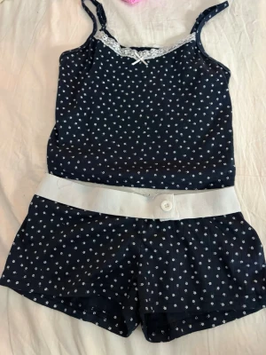Mörkblått pyjamasset med spetsdetalj - Sött tvådelat pyjamasset i mörkblått med små vita blommor. Linnet har smala axelband, spetskant och en liten rosett framtill. Shortsen har bred vit midjeresår med knapp. Perfekt för mysiga kvällar hemma.