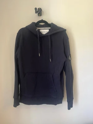 Mörkblå hoodie från Calvin Klein - Snygg mörkblå hoodie från Calvin Klein Jeans med klassisk huva och dragsnören. Stor magficka framtill och CK-logga på vänster ärm. Perfekt för en chill och avslappnad stil. Mjuk bomullskänsla och ribbade muddar vid ärmslut och nederkant. Storlek S, Skick 7/10