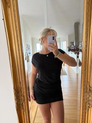 Svart kort klänning med drapering - Snygg svart kort klänning med draperad effekt i midjan och lätt asymmetrisk halsringning. Klänningen är ärmlös med en figurnära passform och tillverkad i ett mjukt, stretchigt material som ger en bekväm känsla.