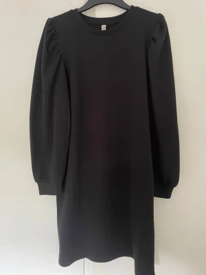 Svart långärmad klänning - En enkel svart klänning med rund halsringning och långa puffärmar. Klänningen har en rak passform och är tillverkad i ett mjukt, stretchigt material som ger en bekväm känsla. Perfekt för dig som gillar minimalistisk stil.