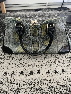 Grön handväska i ormskinnsmönster - Snygg handväska med grönt och svart ormskinnsmönster, svarta lackdetaljer och dubbla handtag. Väskan har guldfärgade metalldetaljer och en dragkedja på baksidan. Perfekt för dig som vill sticka ut med en trendig accessoar.