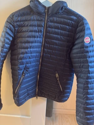 Mörkblå dunjacka från Colmar - Snygg mörkblå dunjacka från Colmar med quiltad design och två dragkedjeförsedda fickor framtill. Jackan har en hög krage och Colmar-logga på ärmen. Perfekt för kalla vinterdagar och riktigt lätt att bära.