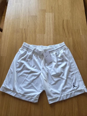 Nike Jordan basket vita shorts, NBA , Dam storlek L - Snygga vita shorts från Jordan med elastisk midja och snörning. Tillverkade i ett lätt och sportigt polyestermaterial. Diskret svart Jumpman-logo broderad på ena benet och liten slits i benslutet för extra rörelsefrihet.