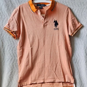 Persikofärgad pikétröja US Polo Assn - Persikofärgad pikétröja från U.S. Polo Assn. med slim fit-passform. Tröjan har orange krage och ärmslut med vita och svarta detaljer, samt broderad logotyp på bröstet. Tillverkad i 100% bomull och har klassisk knappslå framtill.