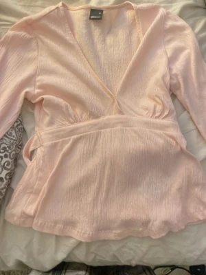 Ljusrosa omlottblus från Gina Tricot storlek Xs - Supersöt ljusrosa omlottblus från Gina Tricot med lång ärm och knytband i midjan. Blusen har en djup v-ringning och är gjord i ett lätt, krinklat tyg som ger en snygg struktur .