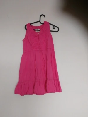 Ärmös klänning i starkt rosa - Snygg ärmlös klänning i starkt rosa med volangkant nertill och knappar framtill. Klänningen har en lätt rynkad struktur och v-ringning. Perfekt för dig som gillar färg och vill sticka ut lite extra.