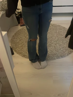 Blå slitna bootcut jeans storlek 30 - Säljer ett par blå bootcut jeans med slitningar och hål på båda benen. Jeansen har klassisk femficksdesign och medel midja .