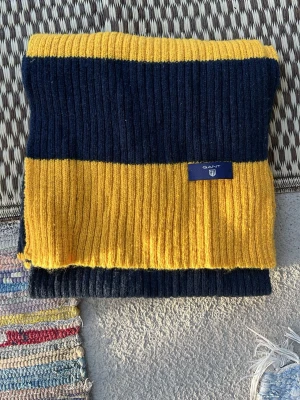 Randig halsduk från Gant i ullmix - Snygg ribbstickad halsduk från Gant i mörkblått och gult. Halsduken har breda ränder och en liten tygpatch med Gant-loggan. Perfekt accessoar för att lyfta din outfit med lite färg.