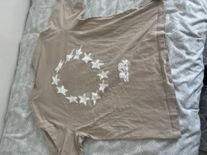 Beige t-shirt med stjärnor och tryck - Beige t-shirt med vita stjärnor i mönster både fram och bak samt trycket 'S/S 202X' på bröstet. T-shirten har klassisk passform och är kortärmad, tillverkad i mjuk bomull. Perfekt för en avslappnad och trendig look.