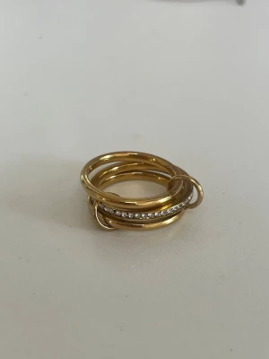 ring  - Säljer en snygg guldfärgad stapelring med flera tunna ringar som sitter ihop. En av ringarna är dekorerad med små gnistrande stenar som ger extra bling. Perfekt för dig som gillar trendiga accessoarer och vill ha något unikt på fingret.