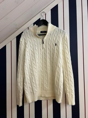   Ralph Lauren Cable knit quarter zip  - Kabelstickad vit tröja från Polo Ralph Lauren med halv dragkedja och hög krage. Tröjan har det klassiska blå broderade logotypen på bröstet och ribbade muddar vid ärmslut och nederkant. Perfekt för lager på lager och en clean look. Den är inte så tjock så är perfekt nu till sommaren. Lite mer åt ”cream-white”  hållet än kritvit