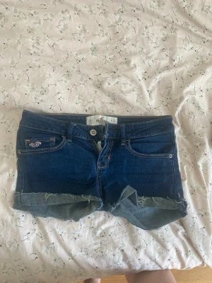 Mörkblå jeansshorts från Hollister - Snygga mörkblå jeansshorts från Hollister med råa kanter och klassisk femficksdesign. Shortsen har låg midja och detaljerade bakfickor med kontrastsömmar. Perfekta för varma dagar och en avslappnad stil. Man kan  justera hur korta man vill att shortsen ska sitta