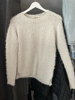 Vit fluffig stickad tröja - Mysig vit stickad tröja med fluffig textur och rund hals. Långärmad modell som är perfekt för kyliga dagar. Tröjan har en avslappnad passform och är tillverkad i ett mjukt material som känns skönt mot huden. 