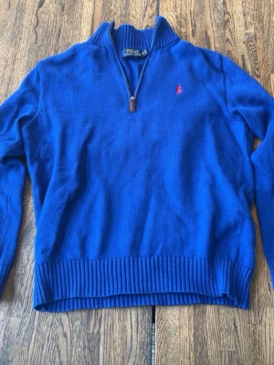 Blå halvzip-tröja från Polo Ralph Lauren - Klassisk blå stickad tröja från Polo Ralph Lauren med halv dragkedja och ribbade muddar. Tröjan har en liten röd logga broderad på bröstet och hög krage. Perfekt för en stilren och avslappnad look.