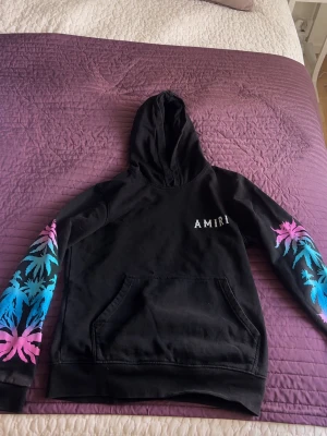 Svart hoodie från Amiri med tryck - Svart hoodie från Amiri med huva och känguruficka. Ärmarna har färgglada palmtryck i blått och rosa, och på bröstet finns en vit Amiri-logga. Perfekt för dig som vill sticka ut med en cool streetwear-look.