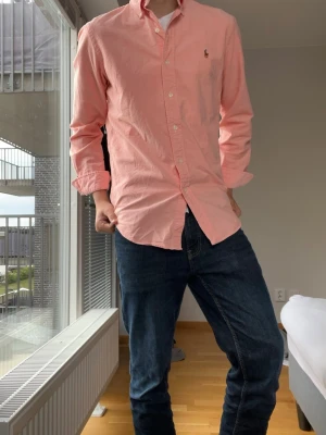 Ljusrosa skjorta från Ralph Lauren - Hallöj! Säljer en ljusrosa skjorta från Ralph Lauren med klassisk button-down krage och broderad logga på bröstet. Skjortan har lång ärm, bra passform och är tillverkad i mjuk bomull. Perfekt för en clean och stilren look. Jag är 178 och väger 66kg och på mig är den lite stor. Jag skulle säga att den passar dig mellan 178-188 ungefär.