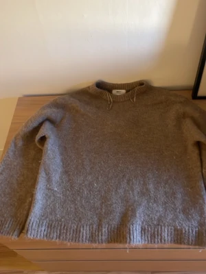 Brun stickad tröja i ull - Mysig brun stickad tröja med rund hals och ribbade muddar. Tröjan har en avslappnad passform och är tillverkad i ett ulligt material som känns både varmt och skönt. Perfekt för kyliga dagar och enkel att matcha med jeans eller kjol.