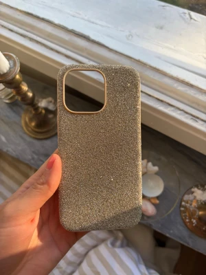 Glittrigt guldigt Mobilskal iPhone 13 Pro Swarovski - Snyggt mobilskal i guld med glittrig yta som verkligen fångar ljuset. Skalets form är anpassad för en modern smartphone med stor kameraöppning. Materialet känns tåligt och skalet har rundade hörn för extra skydd. Perfekt för dig som vill ha ett skal som sticker ut.