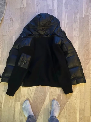 Svart cardiganjacka från C.P. Company - Säljer en svart cardiganjacka från C.P. Company med huva och ribbade muddar. Jackan har en cool ficka med knappdetalj på ärmen och är tillverkad i polyester. Perfekt för kalla dagar när du vill hålla stilen.