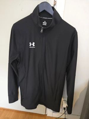Svart ziptröja från Under Armour - Svart ziptröja från Under Armour med dragkedja framtill och hög krage. Jackan har vit logotyp på bröstet och är tillverkad i ett stretchigt, funktionsmaterial som passar perfekt för träning och sportaktiviteter.